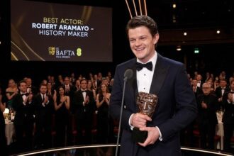 Actor Robert Aramayo smiling widely while holding two BAFTA trophies at the 2026 awards ceremony.