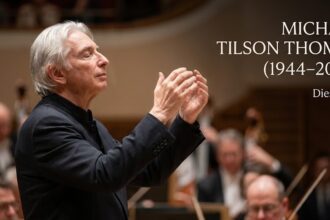 A distinguished portrait of conductor Michael Tilson Thomas passionately leading an orchestra from the podium, baton in hand, bathed in warm stage lighting.